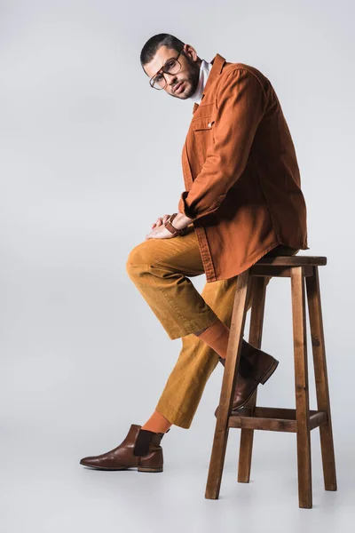 Hombre con gafas y ropa casual mirando cámara cerca de silla de madera sobre fondo gris — Stock Photo