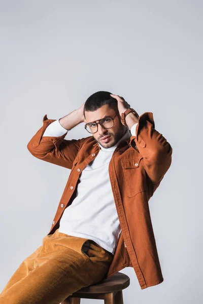 Barbudo hombre con estilo con las manos cerca de la cabeza posando en silla aislada en gris - foto de stock