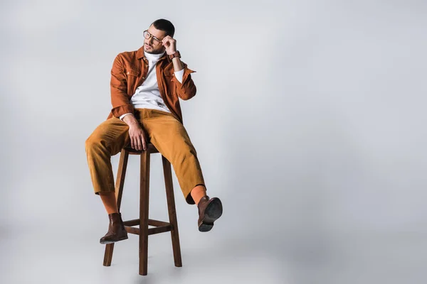 Стильный мужчина с рукой возле очков сидит на деревянном стуле на сером фоне — стоковое фото