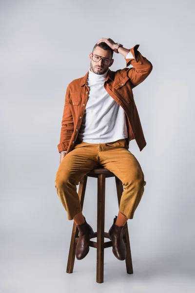 Homme à la mode en col roulé et veste en terre cuite regardant caméra sur chaise en bois sur fond gris — Photo de stock