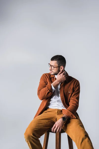 Молодий чоловік у повсякденному одязі та окулярах дивиться на стілець ізольовано на сірому — стокове фото