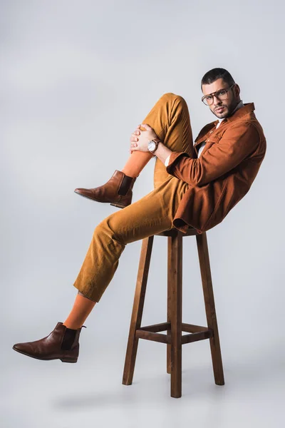 Stylish man in brown shoes and terracotta jacket looking at camera on chair on grey background — Stock Photo