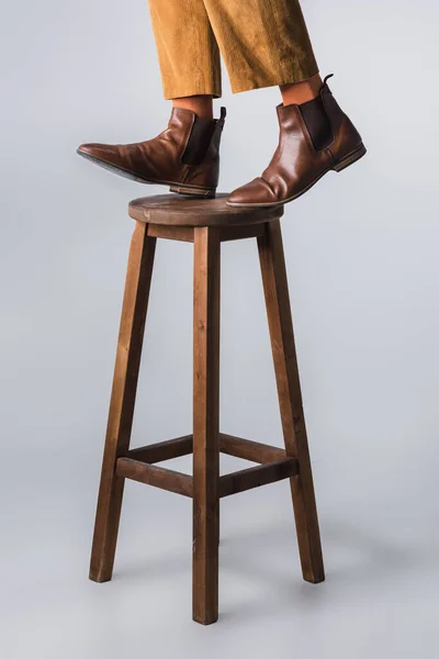 Обрізаний вид людини в коричневому взутті, що стоїть на дерев'яному стільці на сірому фоні — стокове фото