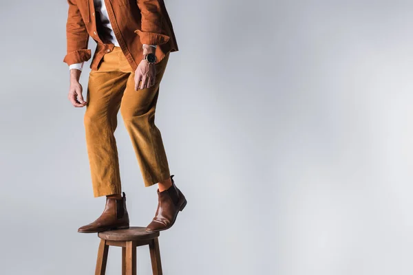 Vista recortada del hombre de moda en zapatos marrones de pie en silla de madera sobre fondo gris - foto de stock
