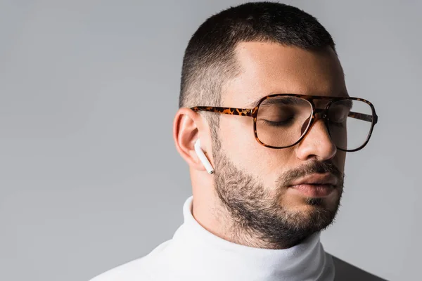 Joven con anteojos y cuello alto escuchando música en auriculares inalámbricos aislados en gris - foto de stock