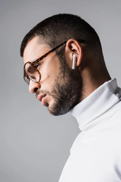 Stylish man in eyeglasses and white turtleneck listening music in wireless earphone isolated on grey — Stock Photo