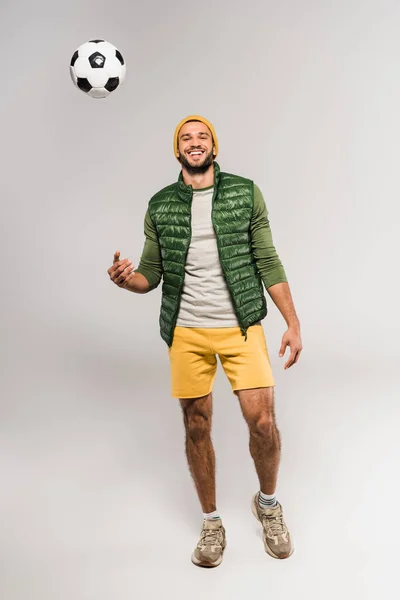 Deportista sonriente mirando a la cámara cerca del fútbol en el aire sobre fondo gris - foto de stock