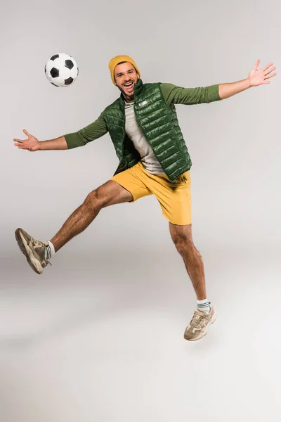 Homem positivo pulando perto de futebol no ar em fundo cinza — Fotografia de Stock