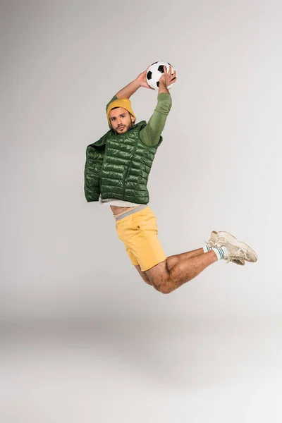 Молодой спортсмен держит футбол, прыгая и глядя в камеру на сером фоне — стоковое фото