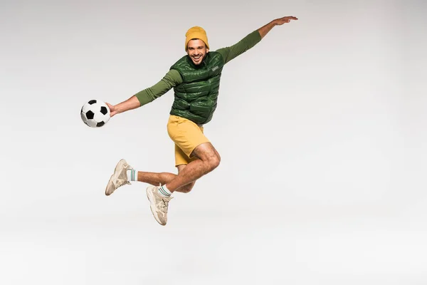 Веселий спортсмен в повсякденному одязі стрибає і тримає футбол ізольовано на сірому — стокове фото