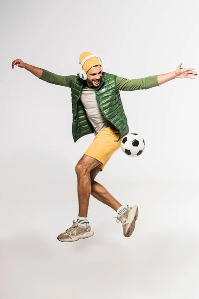 Fröhlicher Sportler mit Kopfhörern, der in Fußballnähe auf grauem Hintergrund springt — Stockfoto