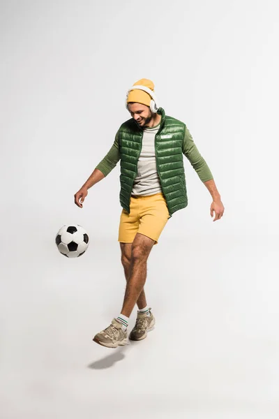 Esportista sorrindo ouvir música em fones de ouvido enquanto joga futebol em fundo cinza — Fotografia de Stock