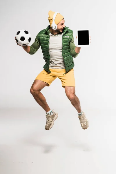 Desportista positivo em fones de ouvido segurando tablet digital com tela em branco e futebol em fundo cinza — Fotografia de Stock