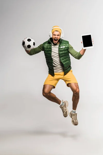 Aufgeregter Sportler mit Kopfhörer, der Fußball und digitales Tablet mit leerem Bildschirm hält, während er auf grauem Hintergrund springt — Stockfoto