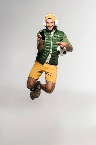 Esportista alegre em fones de ouvido segurando futebol e apontando para o smartphone em fundo cinza — Fotografia de Stock