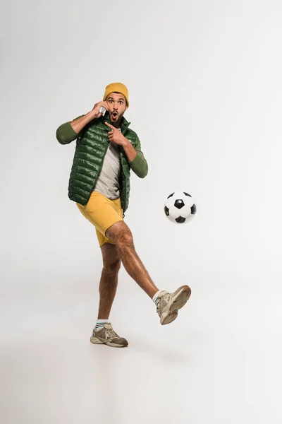 Deportista excitado señalando con el dedo mientras habla en el teléfono inteligente y jugando al fútbol sobre fondo gris - foto de stock
