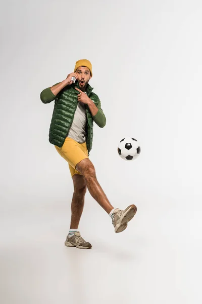 Excited sportsman pointing with finger while talking on smartphone and playing football on grey background — Stock Photo