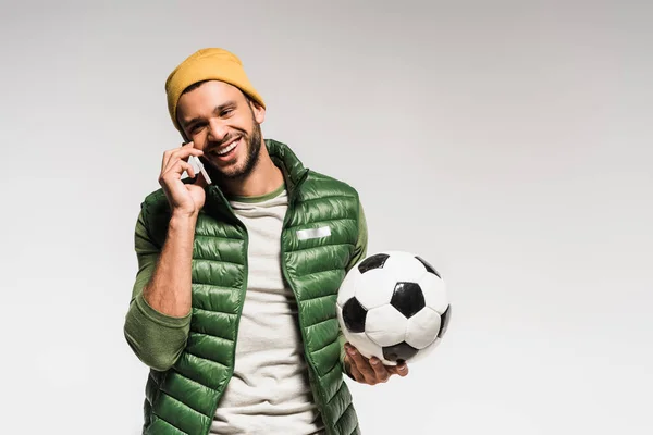 Deportista sonriente sosteniendo el fútbol y hablando en un smartphone sobre fondo gris - foto de stock