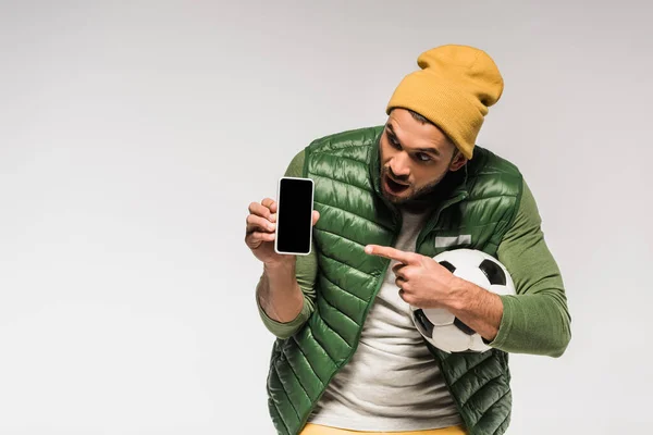 Excited sportsman holding football and pointing at smartphone with blank screen isolated on grey — Stock Photo