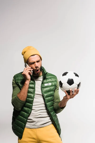 Verblüffter Sportler spricht auf Smartphone und hält Fußball isoliert auf grau — Stockfoto