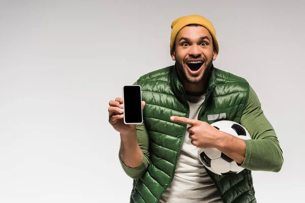 Homem incrível com futebol apontando para smartphone com tela em branco isolado em cinza — Fotografia de Stock