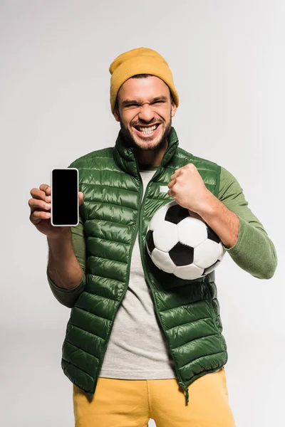 Cheerful sportsman showing yeah gesture and holding smartphone with blank screen and football on grey background — Stock Photo
