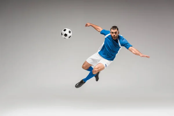 Sportsman olhando para a câmera enquanto saltava perto de futebol no fundo cinza — Fotografia de Stock