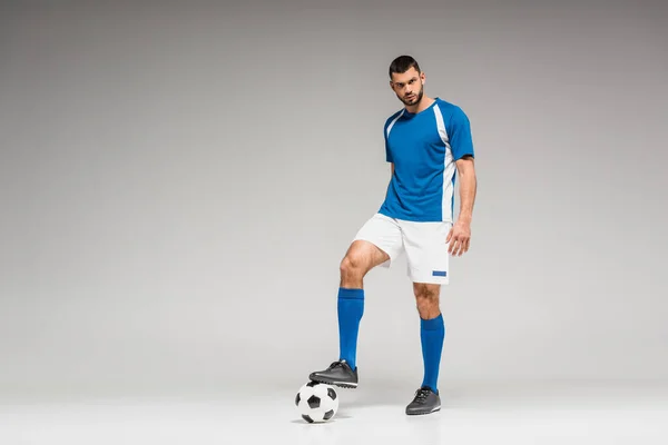 Уверенный спортсмен смотрит в камеру рядом с футболом на сером фоне — стоковое фото