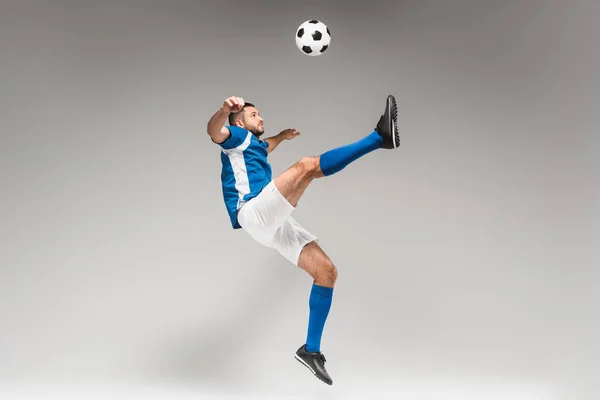 Sportsman in knee socks and sportswear jumping while playing soccer on grey background — Stock Photo