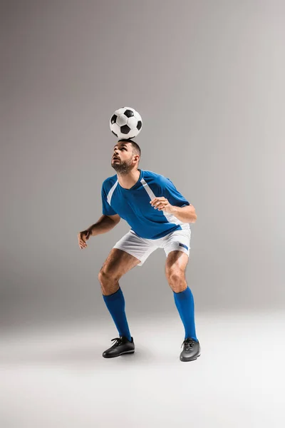 Esportista barbudo equilibrando com futebol na cabeça em fundo cinza — Fotografia de Stock