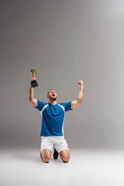 Sportsman with golden champion cup showing yeah gesture while kneeling on grey background — Stock Photo