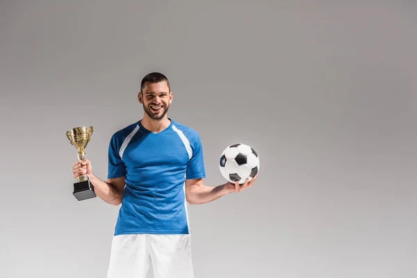 Cheerful sportsman holding football and golden champions cup on grey background — Stock Photo