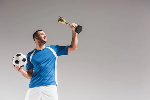 Deportista feliz mirando el trofeo de campeones y sosteniendo el fútbol aislado en gris - foto de stock