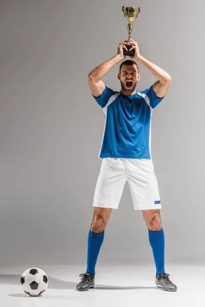 Deportista sorprendido sosteniendo la copa de campeones cerca del fútbol sobre fondo gris - foto de stock