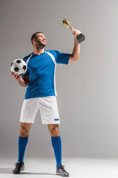 Deportista sonriente sosteniendo el fútbol y mirando el trofeo de campeones sobre fondo gris - foto de stock