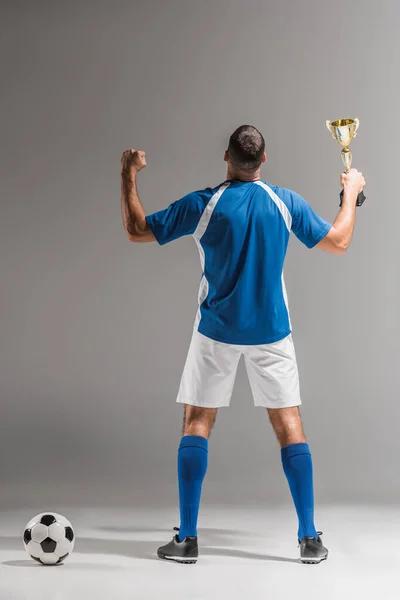 Vue arrière de l'homme athlétique montrant le geste ouais tout en tenant la coupe des champions à côté du football sur fond gris — Photo de stock