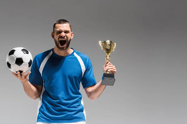 Sportivo stupito con calcio e coppa campioni guardando la fotocamera isolata su grigio — Foto stock