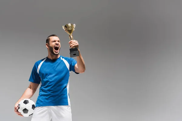 Astonished sportsman looking at golden champions trophy while holding football isolated on grey — Stock Photo