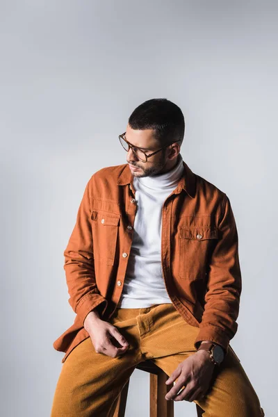 Модный мужчина в пиджаке и очках, сидящий на стуле и отводящий взгляд на сером фоне — стоковое фото