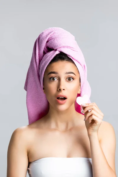 Schockiert schöne Frau mit Handtuch auf dem Haar hält herzförmigen Schwamm isoliert auf grau — Stockfoto