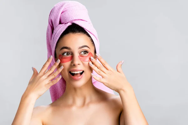 Stupito bella donna con asciugamano sui capelli e macchie oculari idrogel sul viso isolato su grigio — Foto stock