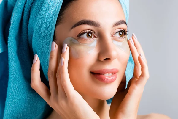 Smiling beautiful woman with blue towel on hair and hydrogel eye patches on face isolated on grey — Stock Photo