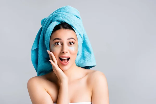 Amazed beautiful woman with blue towel on hair and hydrogel eye patches on face isolated on grey — Stock Photo