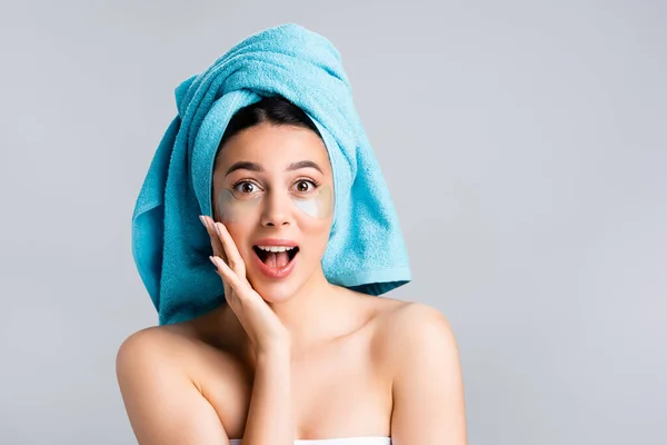 Mujer hermosa asombrada con toalla azul en el cabello y parches de hidrogel en la cara aislada en gris - foto de stock