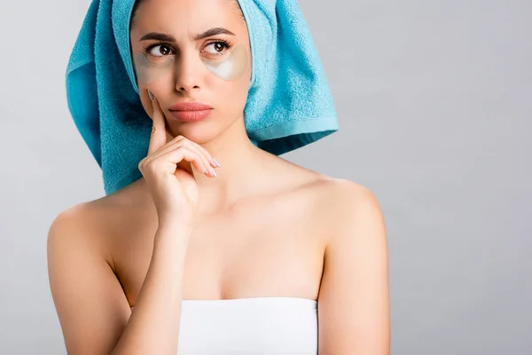 Задумчивая красивая женщина с голубым полотенцем на волосах и гидрогелевые глазные пятна на лице изолированы на сером — стоковое фото