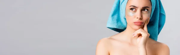 Задумчивая красивая женщина с голубым полотенцем на волосах и гидрогелевые глазные пятна на лице изолированы на сером, баннер — стоковое фото