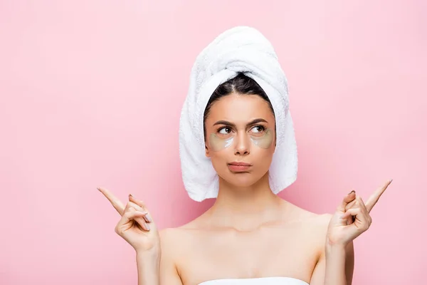 Nachdenklich schöne Frau mit Handtuch auf dem Haar und Hydrogel Augenflecken im Gesicht zeigt mit Fingern isoliert auf rosa — Stockfoto