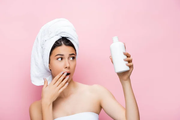 Shocked beautiful woman with towel on hair and bottle of lotion isolated on pink — Stock Photo