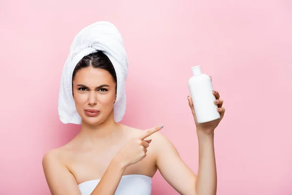 Disgusted beautiful woman with towel on hair pointing at bottle of lotion isolated on pink — Stock Photo