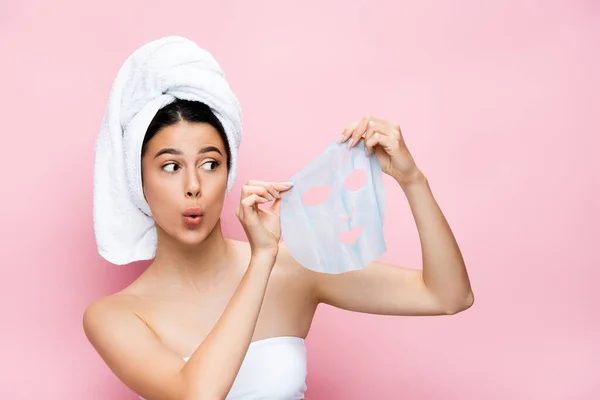 Удивлена красивая женщина с полотенцем на голове держа маску лист изолирован на розовый — стоковое фото